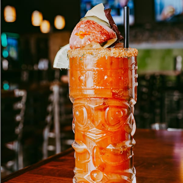 A Tito's Lounge Bloody Mary in a tiki glass, garnished with salami, cheese, and pickles.