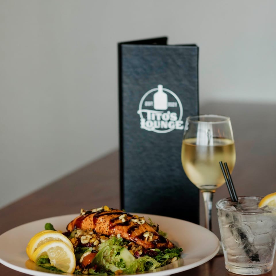 Grilled salmon salad with lemon, a glass of white wine, and a Tito's Lounge menu.