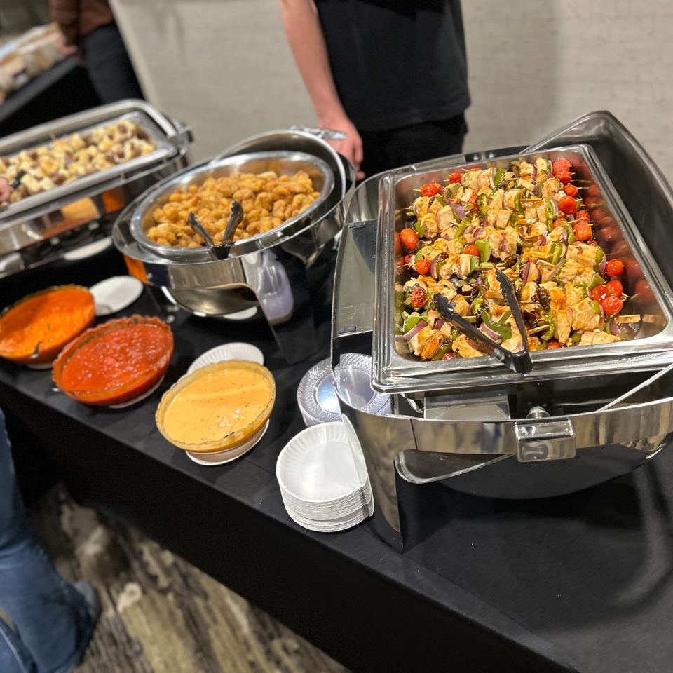 A buffet spread with chafing dishes of chicken skewers, fried food, and bowls of sauces.
