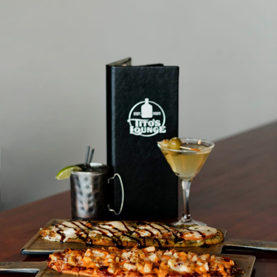 Two flatbread pizzas, a martini, and a Moscow Mule with a Tito's Lounge menu on a wooden table.