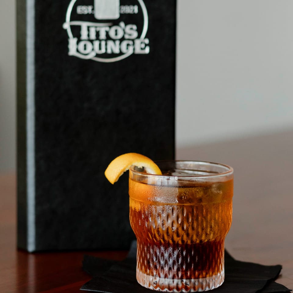 A Tito's Lounge menu stands behind a cocktail with an orange peel on a dark table.